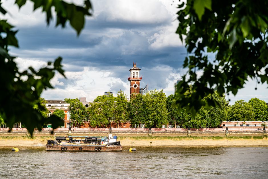 A scenic view of the River Thames with a barge floating along the water under a partly cloudy sky. In the background, there is a historic red-brick clock tower and surrounding greenery including matur