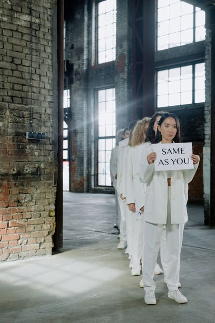 A line of women dressed in white suits standing indoors in front of large industrial windows, with the woman at the front holding a sign that reads 'SAME AS YOU'. The environment features exposed bric