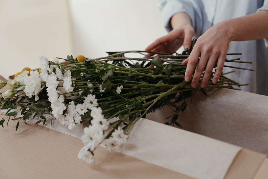 A person’s hands are carefully arranging a bouquet of fresh white carnations, interspersed with sprigs of greenery, on a light wooden surface. The bouquet features a mix of white carnations with ruf
