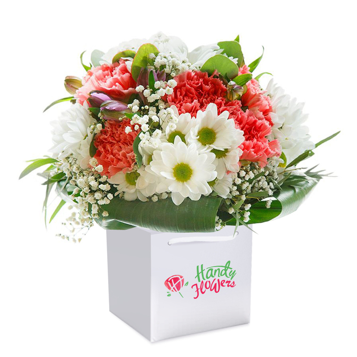 A beautifully curated floral display, drawing inspiration from the vibrant scenes along Whitechapel Road, is showcased upright in a sleek white square gift bag. The focal point is an enchanting mix of softly coloured coral and peach carnations-each with intricate, ruffled petals that ripple gracefully-paired with vivid red carnations that create an energetic counterpoint. The bouquet's heart is brightened by clusters of dazzling white daisies, each with a luminous lime-yellow core, evoking a sense of optimism akin to the atmosphere in Altab Ali Park during summer. Wispy baby's breath interlaces delicately among the blooms, their tiny blossoms resembling flecks of light, while a few purple-hued buds peek through from the side, bringing hints of depth and unfolding possibility. Lush, deep green leaves and glossy foliage embrace the bouquet, shaping its rounded silhouette and grounding the display with the freshness reminiscent of the Columbia Road Flower Market. The arrangement sits proudly within a white gift bag with chic ribbon handles for effortless carrying, embodying both style and practicality for celebrations or surprise deliveries throughout Whitechapel and beyond. A pristine white backdrop ensures that every colour, contour, and artisan detail stands out clearly-demonstrating the quality and vibrancy Whitechapel Florist brings to every creation. This bouquet not only celebrates the everyday beauty found throughout East London but also serves as a perfect gesture for anniversaries, birthdays, or any occasion that calls for heartfelt flowers.